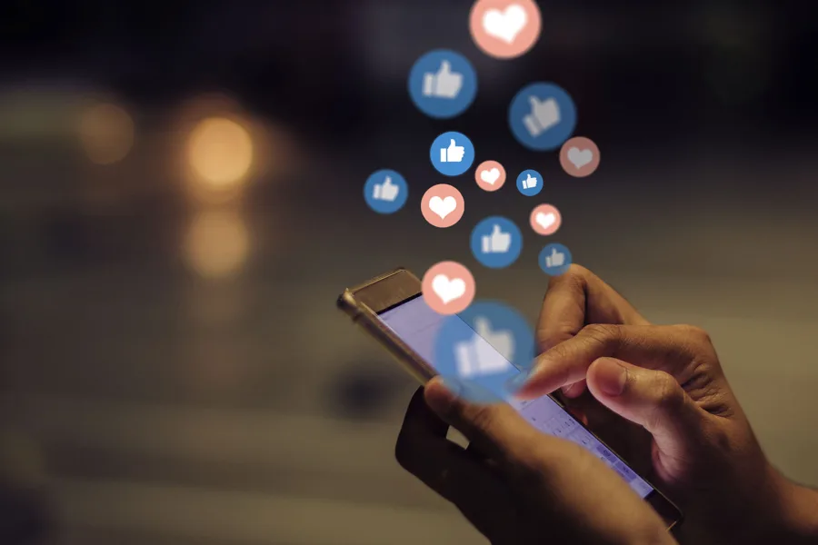A person holding tapping a mobile phone with emojis floating out of it.